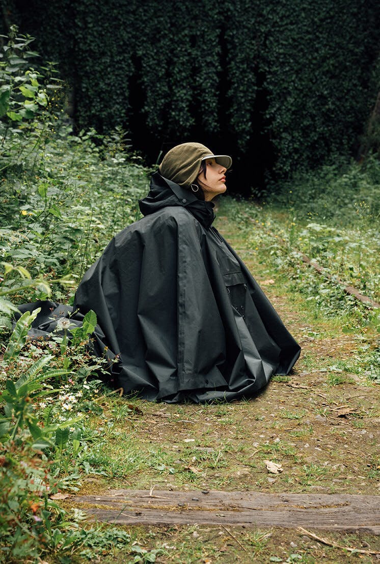 Person in a dark green cape sitting in a lush green abandoned train track.
